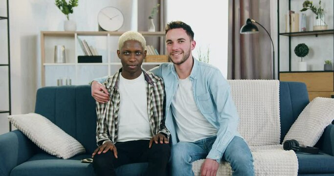 Portrait Of Attractive Happy Smiling Mixed Race Male Couple With Unconventional Sexual Orientation Which Looking Into Camera Sitting On The Couch At Home,one Guy Hugging His Partner By The Shoulder