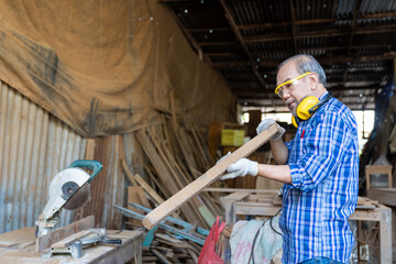 Asian senior male carpenter examining piece of wood in carpentry workshop. Carpenter working his job at the workshop