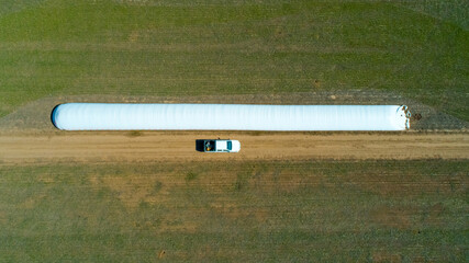 Aerial view of 4x4 pickup truck driving through wheat crops field with silos bags on the road....