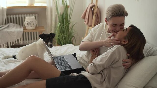 Lesbian women using laptop and kissing while lying on bed at home interior spbd.