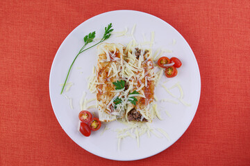 Pancakes with meat, in tomato sauce and cheese, with cilantro and grated cheese on top, and some cherry tomatoes on the plate. Top view on red background.