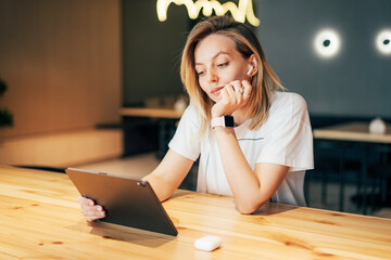 A young attractive blonde sitting at a table in a cafe looks at the screen and listens to audio
