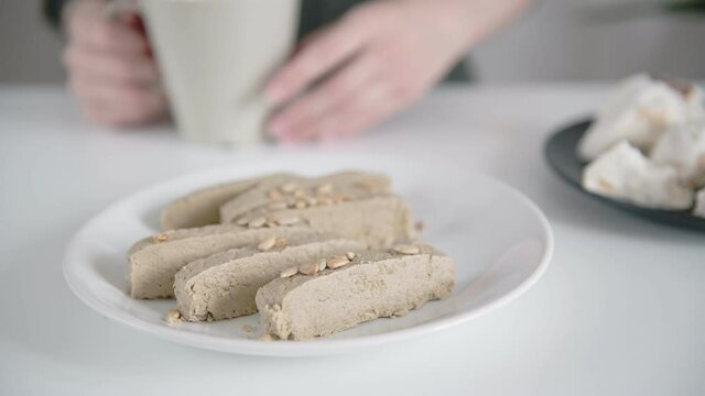 Blurred young woman in shirt drinks tea from white cup and eats brown halva slice taking from plate on foreground close view