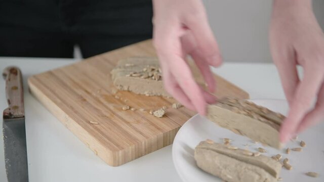 Lady cook serves brown halva slices taking from wooden board and putting on white plate with knife on kitchen table close view.