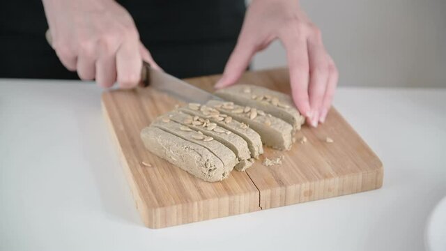 Young woman hands cut brown halva into slices on wooden cutting board with sharp steel knife on kitchen table close view.