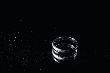 One silver ring without a precious stone on black background with a mirror surface with drops of splashes. Copy space. Platinum jewelry. White gold wedding symbol. Metallic object. Concept of divorce