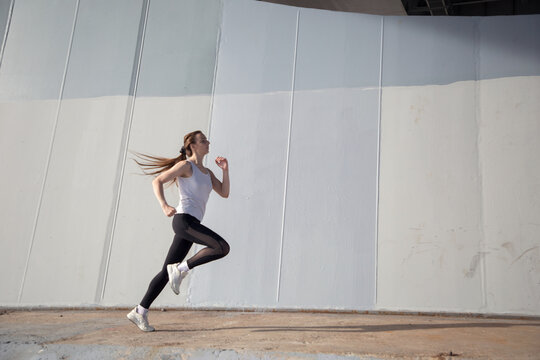 A young woman in a white T-shirt and black pants runs under the bridge in the evening. Running trainings in the city. Preparing for the competition.