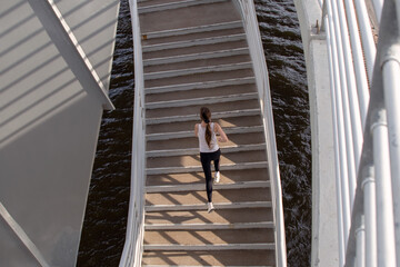 Beautiful top view of the athlete girl running on the stairs of the bridge. Evening strength running workout in the city. Preparing for the competition.