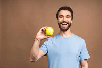 Diet concept. Cheerful beautiful bearded young man eating green apple, isolated over beige background.