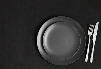 Black table setting. Empty black plate and silver cutlery on a dark concrete background. View from above. Copy space.