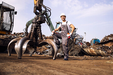 Portrait of junkyard worker standing by hydraulic industrial machine with claw attachment used for lifting scrap metal parts.