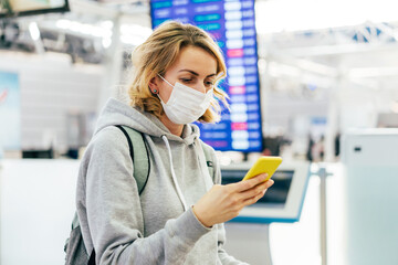 Woman in a medical mask with a mobile phone at the airport near the electronic board of departure.