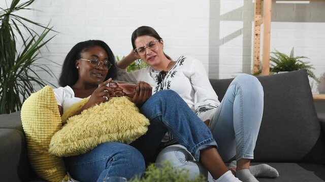 Two young woman black African and white couple in lesbian relationship sitting at home on the sofa and having fun watching videos on the smartphone screen and enjoying their free time together