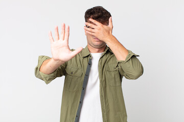 young handsome man covering face with hand and putting other hand up front to stop camera, refusing...