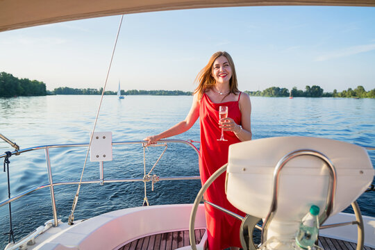 The elegant woman in a long red dress holding a glass of champagne standing on a yacht and looking for river sunset view