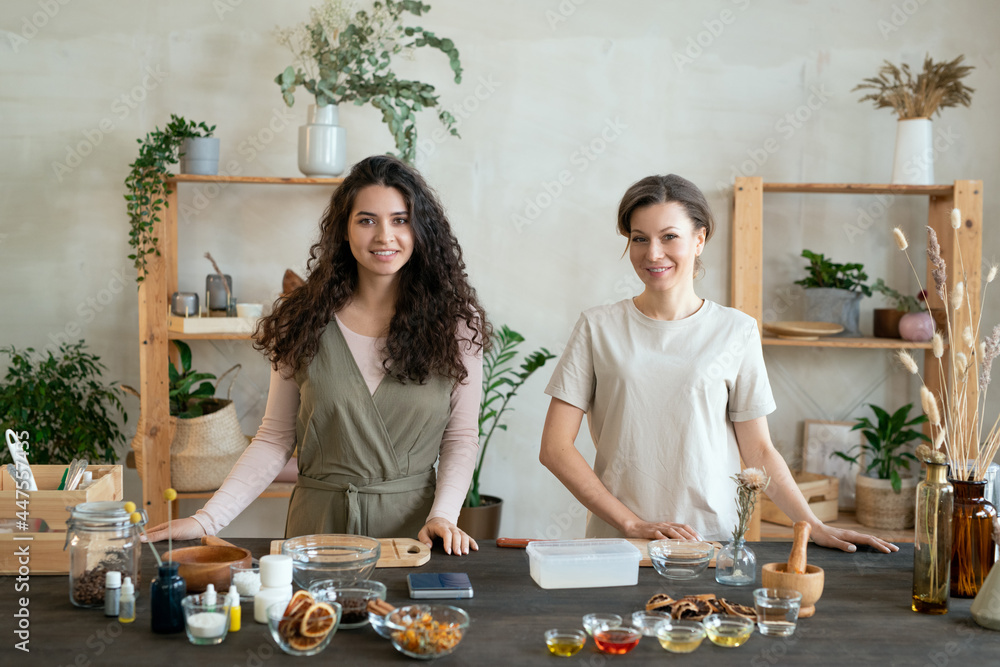 Poster two young females standing by table while one of them cutting hard soap mass