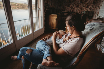 Mother breastfeeding baby on chair in vintage room looking out on lake