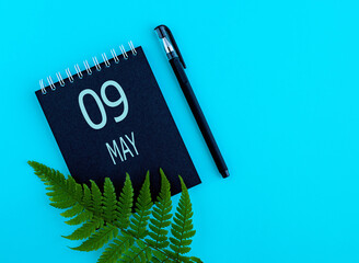 May 9th. Day 9 of month, Calendar date. Black notepad sheet, pen, fern twig, on a blue background....