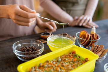 Hands of female pouring melted soap mass into container with dry flowers