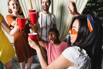 Excited african american friend holding plastic cup near friends in patio