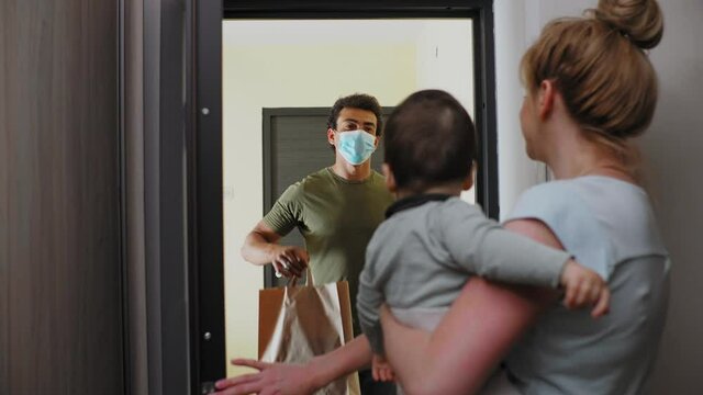 Delivery Man With Face Mask Delivers Package To A Mother With A Baby In Her Arms.