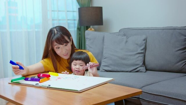 Young mom helping daughter drawing with colored pencils in living room at home.