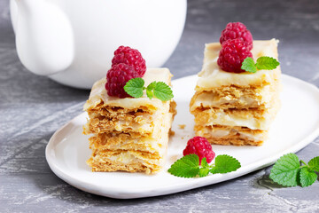 Pieces of Napoleon cake decorated with raspberries and lemon balm leaves on a light background.