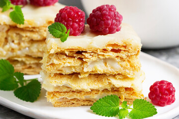 Pieces of Napoleon cake decorated with raspberries and lemon balm leaves on a light background.