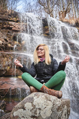 Fototapeta na wymiar Young woman meditating next to a waterfall in a forest, relaxing in warm clothes, enjoying autumn time isolated in nature, all by herself
