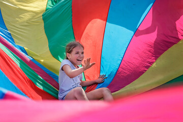Happy girl having fun and waving in the center of bright colorful parachute, outdoors party, summer...