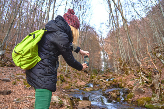 Young woman, a hiker or a backpacker, walking through the forest, taking images and photographs of winter nature, cold river and autumn leaves