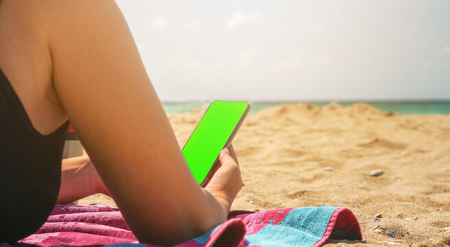 Young female checking on social media on her phone on the beach. . Green screen chroma key smartphone display. Concept of freelancing, working from the beach, vacations, holidays, and remote working. 