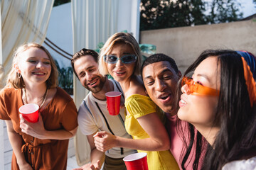 Multicultural friends with plastic cups laughing and looking at asian woman outdoors