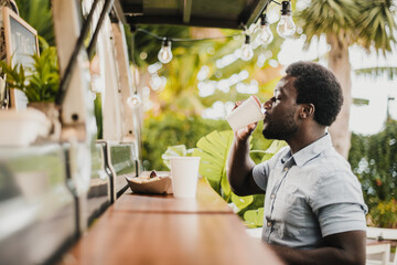 Young african man eating and drinking in food truck counter outdoor in city park - Focus on face.