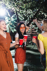 Cheerful interracial women with plastic cups dancing during summer party at night