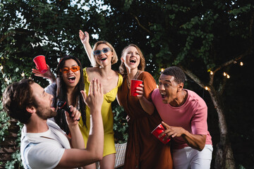 Man singing in microphone near positive multicultural friends with plastic cups outdoors in evening