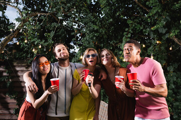 cheerful multiethnic friends holding plastic cups while singing karaoke in garden at night