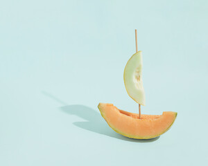 Obraz na płótnie Canvas Sunlit sailing boat made of pale green and orange melon slices and skewer stick, isolated on pastel blue background. Summer vacation sea concept. Minimal holiday, ocean scene. Sun and shadows.