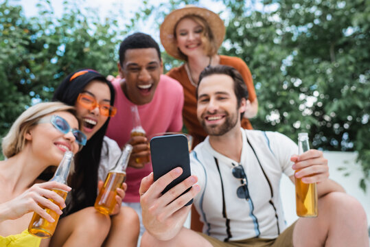 happy man taking selfie with blurred multiethnic friends during beer party
