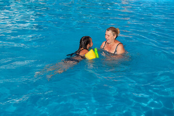 Grandmother and granddaughter in the pool. A kind grandmother teaches her granddaughter to swim in green inflatable sleeves in the summer in the hotel pool on a family trip. 