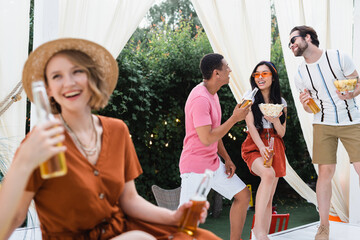blurred woman in straw hat laughing near multiethnic friends drinking beer in patio