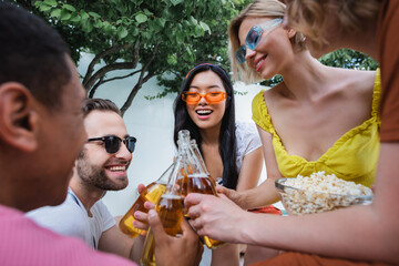 excited multicultural friends clinking bottles of beer during party on blurred foreground