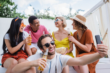 cheerful man in sunglasses taking selfie near interracial friends drinking beer