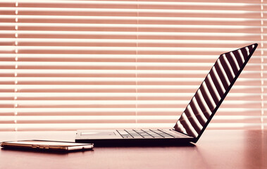 Side view of modern workplace with laptop computer and cellphone liying on table above window with blinds. Warm sunset light.