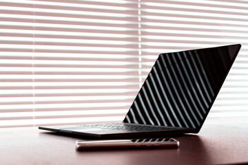 Workplace with opened laptop computer and cellphone liying on table above window with blinds. Natural sunset light with deep shadows.