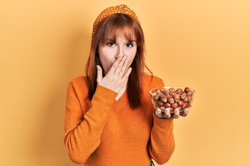 Redhead young woman holding hazelnuts covering mouth with hand, shocked and afraid for mistake....