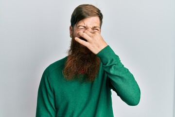 Redhead man with long beard wearing casual clothes smelling something stinky and disgusting,...