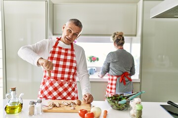 Middle age caucasian couple cooking healthy salad with angry face, negative sign showing dislike with thumbs down, rejection concept