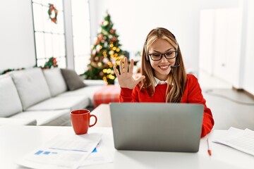 Young caucasian girl sitting on the table working using laptop by christmas tree showing and pointing up with fingers number five while smiling confident and happy.