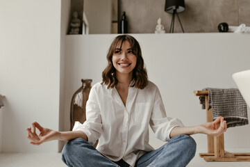 Young happy brunette woman in trendy denim pants and white blouse smiles and meditates. Joyful curly lady sits on floor.